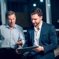 Two office workers with tablet in office late at night. Young businessmen talking while viewing presentation on digital touchscreen tablet.